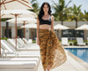 Woman in a black bikini top and tiger print sarong standing by a poolside with palm trees in the background.