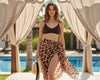 Woman in a brown bikini top and giraffe print sarong standing by a poolside with curtains.