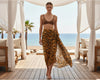Woman in a brown bikini top and tiger print sarong standing on a wooden deck with ocean view.