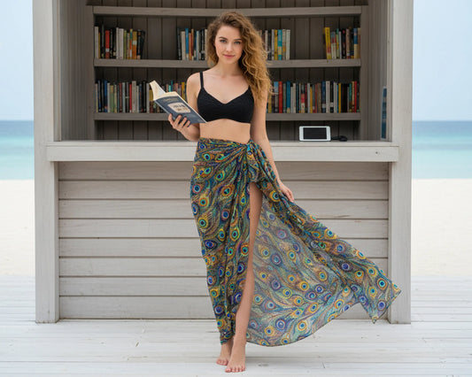Woman holding a book and wearing a colorful patterned sarong in front of a beachside bookshelf.