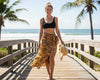 Woman in a black sports bra and tiger print sarong standing on a wooden boardwalk by the beach.